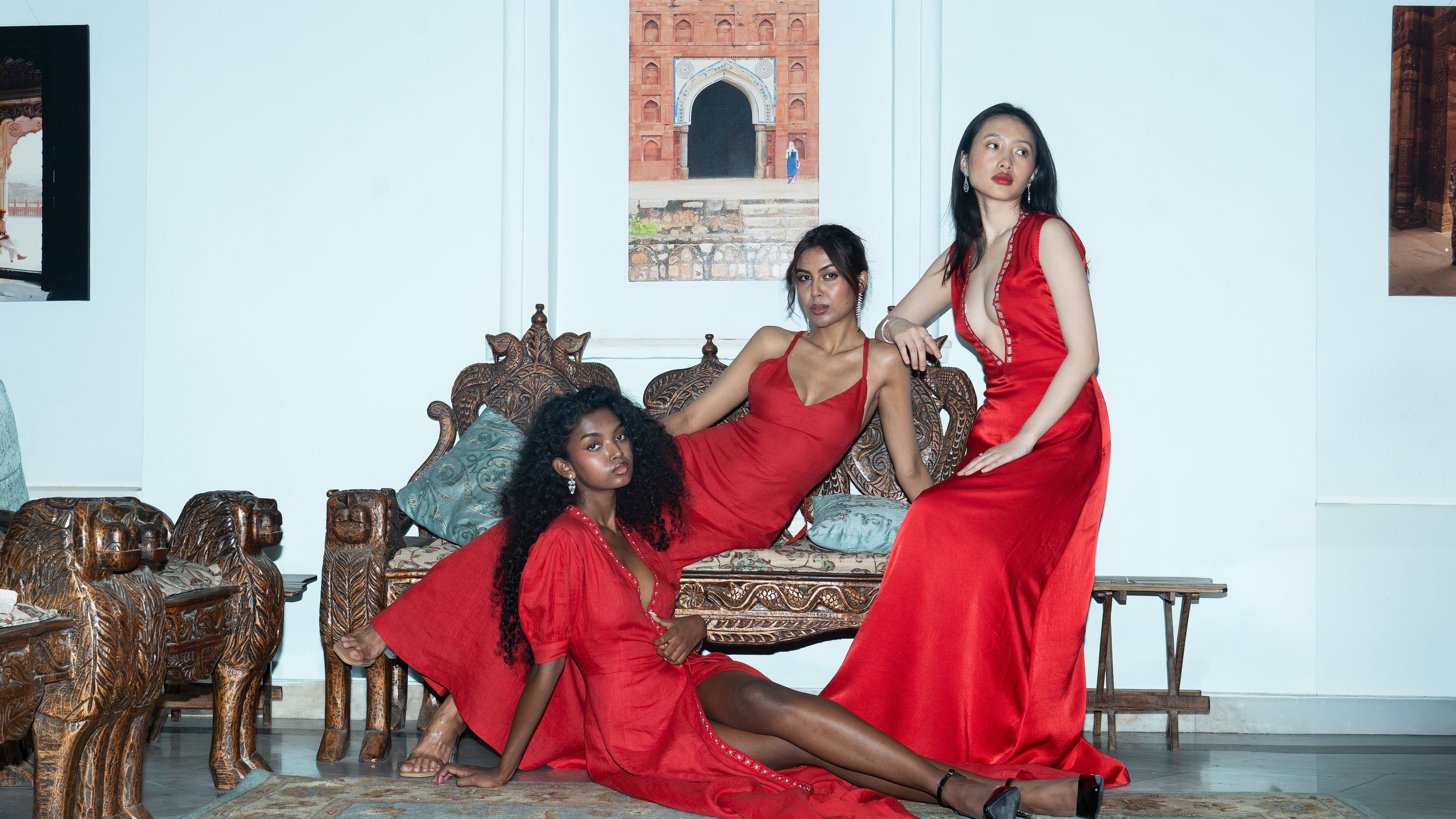 Three women in red dresses sitting on a ornate wooden bench in a room with white walls and decorative elements.
