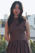 Woman in a brown dress standing outdoors with greenery and buildings in the background