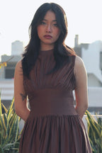Woman in a brown dress standing outdoors with greenery and buildings in the background